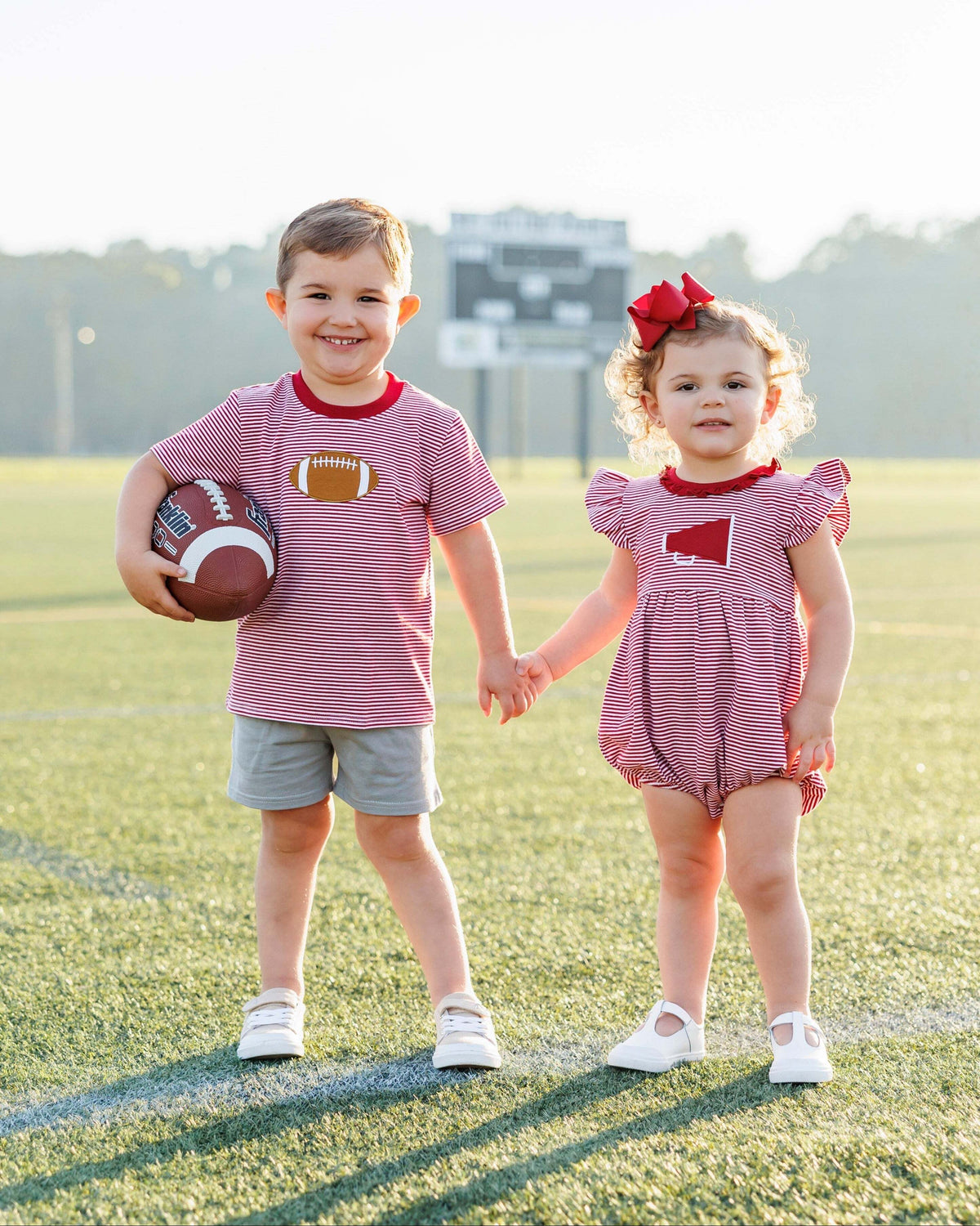 Two children, a boy and a girl, standing on a sports field holding hands. The boy is wearing a red and white striped shirt with a football design and gray shorts, while the girl is in a matching outfit with a red bow.