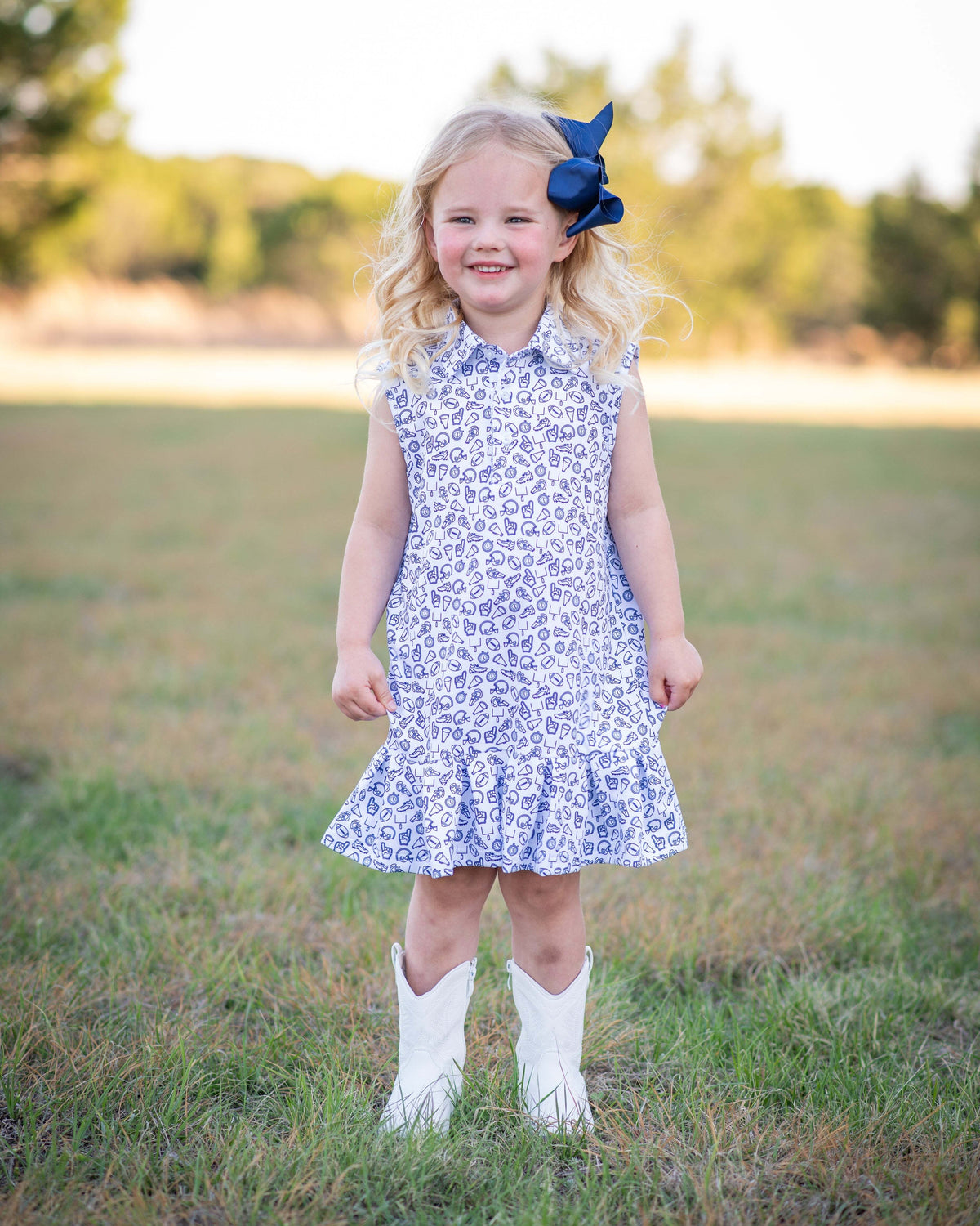 A young girl with blonde curly hair stands in a grassy field wearing a blue and white patterned sleeveless collared dress with a ruffled hem, white cowboy boots, and a navy blue bow in her hair.