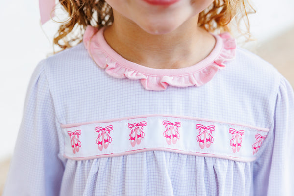Close-up of a child wearing a light lavender gingham smocked dress with a pink ruffled collar and a decorative band featuring pink ballet shoes.