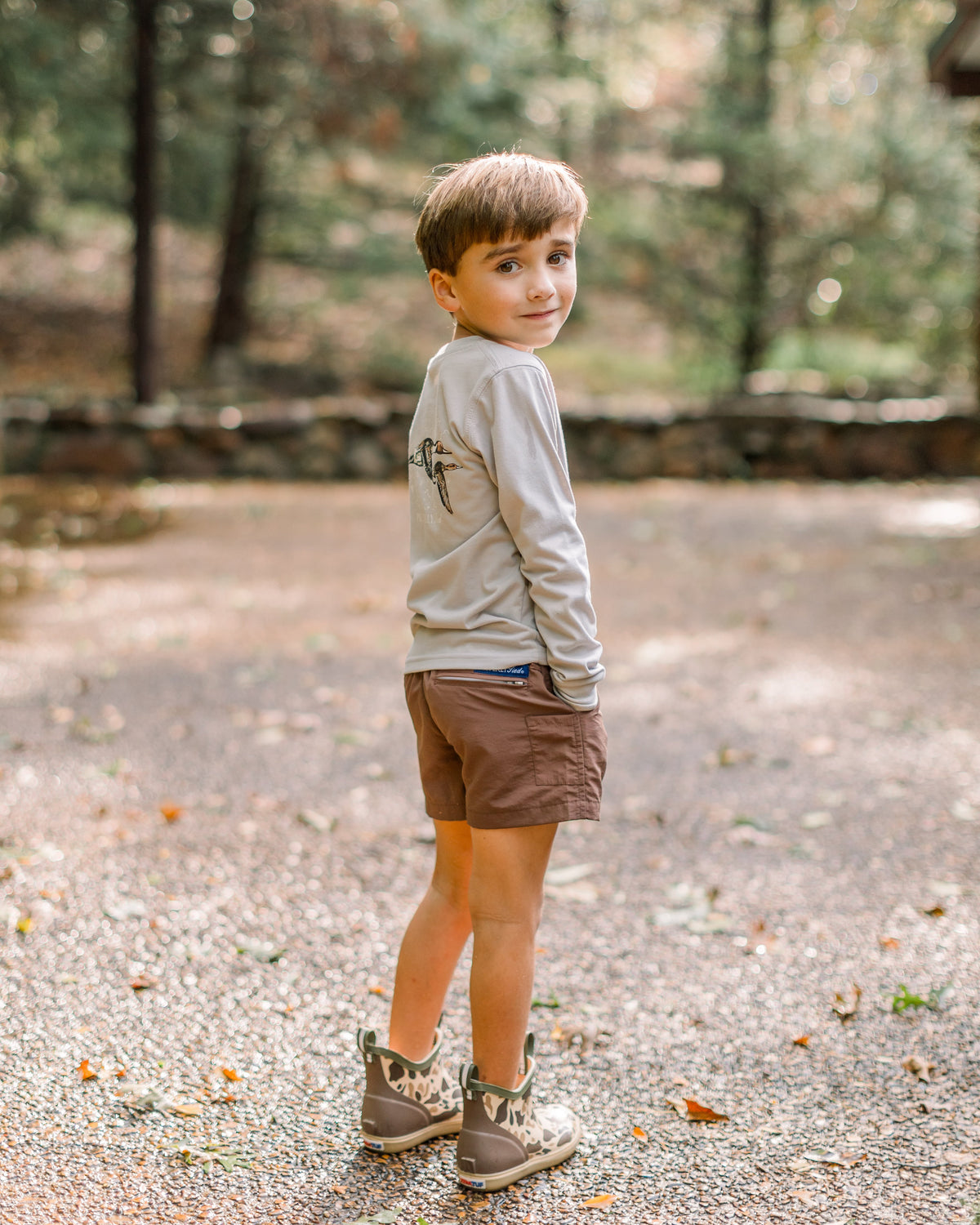 A young boy stands outdoors on a gravel path in a wooded setting, wearing a light gray long-sleeve shirt, brown shorts, and brown-and-gray sneakers.